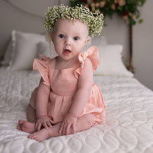 Load image into Gallery viewer, Picture of an infant sitting on a bed with a white bed cover. She has a wreath of small white flowers on her head. She is wearing a peach coloured linen romper with  ruffled short sleeves.