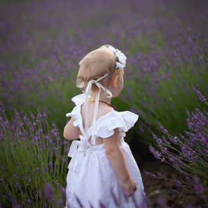 Small light haired child wearing a white rose flower crown, tied with ribbon, and a white linen dress with ruffled sleeves. She's standing in the afternoon summer sun, back facing the camera, in a field of lavender. The criss-cross straps of the dress can be seen.