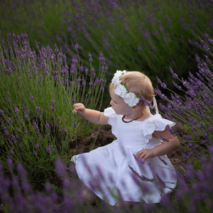 A small fair haired child wearing a white flower crown and white linen dress with ruffle sleeves sits in a field of lavender. She's picking a flower.