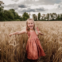 Load image into Gallery viewer, A little girl with long blond hair stands in a wheat field with trees in the distance. She has a big open mouth smile and her arms extended. She is wearing a copper coloured linen dress with ruffle sleeves.