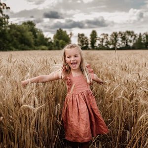 A little girl with long blond hair stands in a wheat field with trees in the distance. She has a big open mouth smile and her arms extended. She is wearing a copper coloured linen dress with ruffle sleeves.